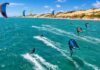 Aerial action photo of kitesurfers and wingfoil riders racing across turquoise ocean water with strong wind-filled kites…