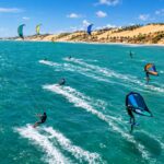 Aerial Action Photo Of Kitesurfers And Wingfoil Riders Racing Across Turquoise Ocean Water With Strong Wind-Filled Kites…