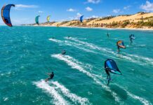 Aerial action photo of kitesurfers and wingfoil riders racing across turquoise ocean water with strong wind-filled kites…