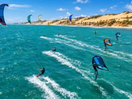 Aerial action photo of kitesurfers and wingfoil riders racing across turquoise ocean water with strong wind-filled kites…