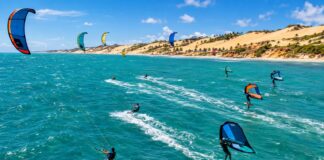 Aerial action photo of kitesurfers and wingfoil riders racing across turquoise ocean water with strong wind-filled kites…