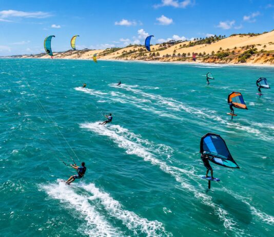 Aerial Action Photo Of Kitesurfers And Wingfoil Riders Racing Across Turquoise Ocean Water With Strong Wind-Filled Kites…