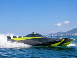Uma lancha catamarã de competição offshore em altíssima velocidade, cortando as águas do litoral brasileiro em pleno dia…