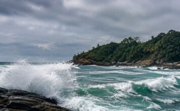 Naufrágio Em Ilhabela Expõe Falhas De Segurança No Mar Foto Realista De Um Mar Agitado Batendo Em Rochas Na Costa De Uma Ilha Tropical Brasileira, Céu Nublado, Sem Pessoas, Se…